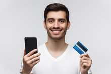Studio Portrait Of Young Smiling Man Wearing White T-shirt, Holding Credit Card And Phone, Isolated On Gray Background