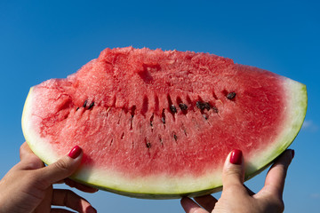 A large slice of watermelon in female hands against the blue sky