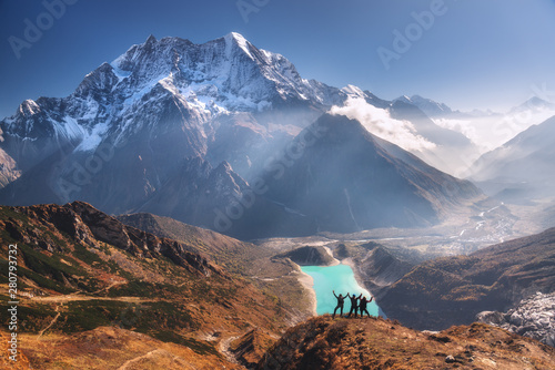 Fototapeta Himalaje  szczesliwi-ludzie-z-podniesionymi-rekami-na-szczyt-przed-pieknym-jeziorem-zasniezona-gora-o-wschodzie-slonca-krajobraz-z-mezczyzna-i-kobieta-skaly-w-chmurach-blekitne-niebo-z-promieniami-slonca-podrozuj-po-nepalu-trekking