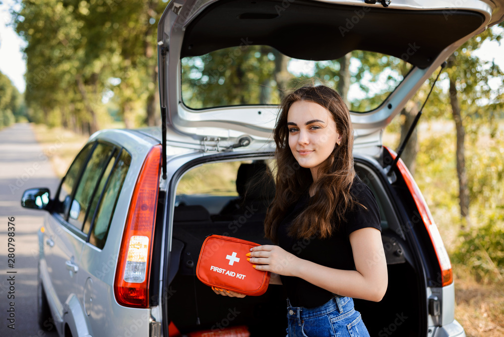 Young girl standing near open back door of silver hatchback car and shows first aid kit that must be in every car for emergency - obrazy, fototapety, plakaty 