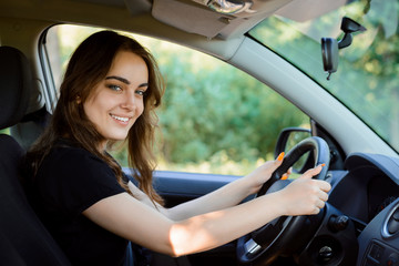 Wall Mural - Attractive cheerful girl driver sits in the driver's seat of a modern car and looks to the camera with broad smile