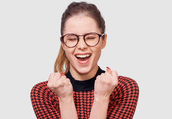 Horizontal studio portrait of young woman with closed eyes, clench fists, rejoices the success, wearing round transparent eyewear, posing over white background.