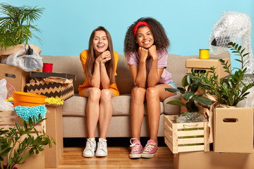 Glad diverse women friends have moving day in new home, relax after cleaning and unpacking, have happy expresisons take rest on sofa in living room rejoice buying house cardboard boxes with belongings