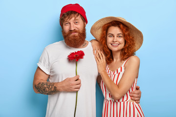 Family portrait of cheerful redhaired husband and wife, cuddle and have caring looks, man holds red gerbera, expresses love to girlfriend in summer hat and striped dress. Relationship, dating concept