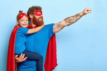 Glad family being heroic leaders, wear superman costumes, have determination to achieve goal and challenging obstacles, caring father holds little girl, ready for flight, isolated on blue background