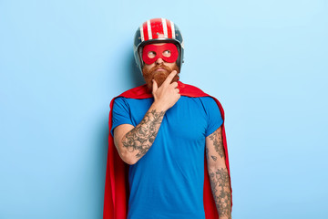 Contemplative young bearded man with tattooed arms, thinks about flight, wears headgear, t shirt, cape and mask, pretends being crime fighting character with supernatural power, poses indoor