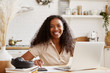 © Anatoliy Karlyuk - Attractive stylish young dark skinned female in beige shirt sitting at kitchen table, using laptop, calculating budget, planning vacations, smiling happily. Self employed black woman working from home