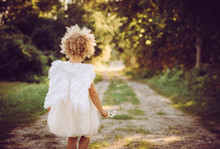 Super Cute Anonymous Blonde Curly Hair Girl Child Wearing White Dress And Angel Wings, Walking On Countryside Road. Backside View. Concept Of Child Angel.