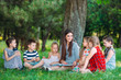 © davit85 - Children and education, young woman at work as educator reading book to boys and girls in park.