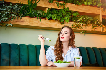 Wall Mural - Young happy woman eating salad in the beautiful interior with green flowers on the background and fresh ingredients on the table. Healthy food concept