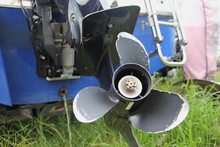 Close Up Lower Unit Of Boat Transom Outboard Motor - 3 Blades Propeller Gear, Bracket Clamp With Trim And Tilt Cylinder On Blue Boat Transom And Green Grass Background