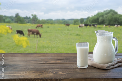 Fotomural Fresh milk in glass on dark wooden table and blurred landscape with cow on meadow