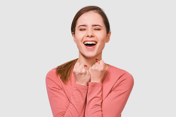 Joyful young woman with closed eyes, clench fists, rejoices the success, wears pink casual long sleeve shirt, poses over white studio background