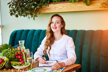 Portrait of a young woman nutritionist in medical gown sitting in the office with healthy food on the background. Weight loss and right nutrition concept