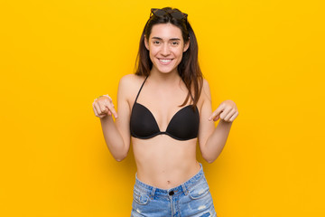 Wall Mural - Young brunette woman wearing a bikini against yellow background points down with fingers, positive feeling.