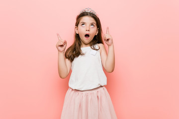 little girl wearing a princess look pointing upside with opened mouth.
