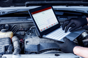 Mechanic is repairing car at service station. Closeup laptop screen with special software at vehicle with open hood. Repairman is conducting diagnostics and detecting problems at car repair shop.