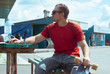 A young hipster man in a red t-shirt and sunglasses resting in a place for camping, traveling on a longboard,
