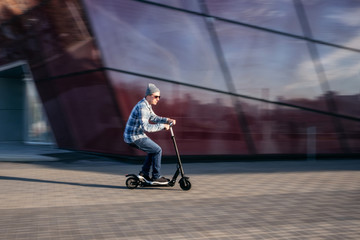 Wall Mural - Young man in casual wear on electric kick scooter on city street in motion blur against modern glass office building