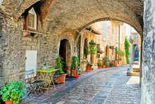 Beautiful Arched Street In The Medieval Old Town Of Assisi With Flowers And Restaurant Tables, Italy