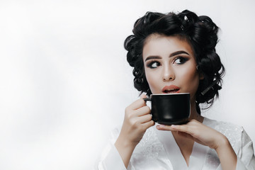 Wall Mural - Lovely ordinary girl in hair curlers and bathrobe, holding cup while drinking coffee or tea in morning. Good morning. She enjoys a hot drink.