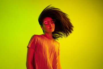 Wall Mural - Beautiful female half-length portrait isolated on yellow studio background in neon light. Young emotional woman. Human emotions, facial expression concept. Dance in eyeglasses and wireless headphones.