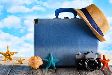 Blue suitcase for summer holidays, hipster hat; retro photo camera and shells on wooden table and beach