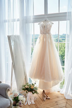 Beautiful Beige Wedding Dress Hanging On Hanger Against Window In Hotel Room, Copy Space. Bridal Bouquet And Women's Shoes Standing On Wood Floor