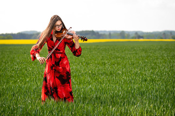 Young woman in red dress playing violin in green meadow - image