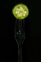 Slice Of Fresh Green Cucumber On Fork On Dark Black Background. Silhouette Of A Fork In The Back Light