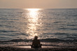© zoranlino - Girl sitting on sandy beach next to sea and enjoys watching sunset, reflection of sun rays over sea. Silhouette of woman by ocean. Light and shadow, woman rests during sunset at tropical landscape.
