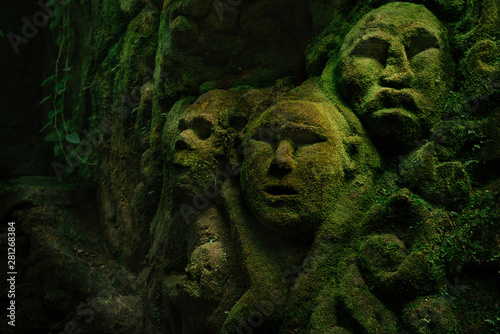 Fotografía Carving demons faces on wall background covered with moss texture in Bali