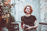 Fototapeta  - Young hipster female journalist with retro camera  sits on a chair and smiles outdoors against the backdrop of garlands