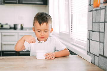 Wall Mural - Cute boy in the kitchen eating yogurt from a white yogurt container, a place for advertising