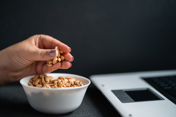 Unhealthy snack at workplace, salt amount. Hands of woman working at computer and taking peanuts from the bowl. Bad habits, salted food, high calorie eating, weight gain and lifestyle concept