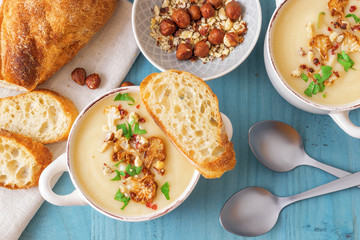 Blue wooden table with 2 bowls of cauliflower soup embellished with crushed hazelnuts, red pepper, smooth parsley herbs, Swiss twisted bread, frying pan with cauliflower, mortar and yellow flowers.