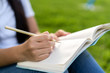Close up of a female hand writing something on the paper green grass background.women sitting relaxed use pencil and notebook on a green grass in college.