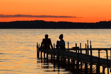 Summer nature background with sunset on the lake.Scenic view with silhouettes of man and woman enjoying beautiful sunset on the wooden pier on the lake.Bright color after sunset sky reflects in water.