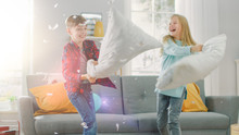 Adorable Little Boy And Sweet Little Girl Have A Pillow Fight In The Sunny Living Room. Siblings Having Fun Fighting With Pillows, Feathers Flying Around.