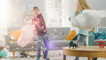 Adorable Little Boy And Sweet Little Girl Have A Pillow Fight In The Sunny Living Room. Siblings Having Fun Fighting With Pillows, Feathers Flying Around.