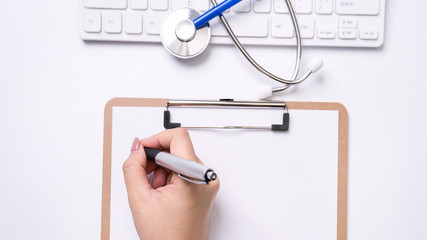 Wall Mural - Female doctor writing a medical record case over clipboard on white working table with stethoscope, computer keyboard. Top view, flat lay, copy space