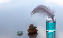 A Bottle Of Blue Scented Oil And A Sticking Bird Feather. A Pyramid Of Stones And A Piece Of Glass Is Folded. Blue Blurred Background. Place For Text.