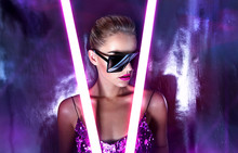 Portrait Of A Beautiful Young Tanned Girl In Big Sunglasses In A Pink Shiny Dress In The Studio On A Silver Background With A Pink And Purple Hue. Glowing Fluorescent Lamps In The Face.