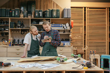 Two Carpenters Craftsmen Are Examining The Drawings Of The Product Of A New Project While Standing In Their Carpentry Against The Background Of Shelves With Tools. Concept Of Hard Work