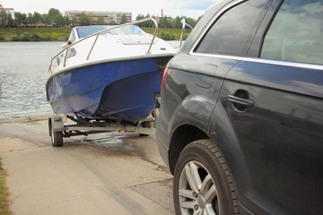 Wall Mural - A 4x4 black car pulls blue-white trimaran cabin motor boat on trailer on concrete slipway into the river water, DIY boat launch