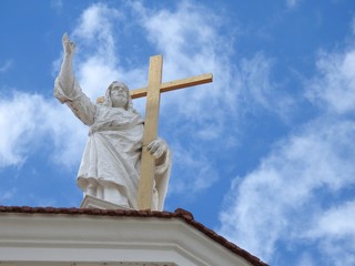 A stone statue of Jesus Christ, with his hand raised and holding a large cross, on top of a church. Sunny day with clear sky and some clouds.