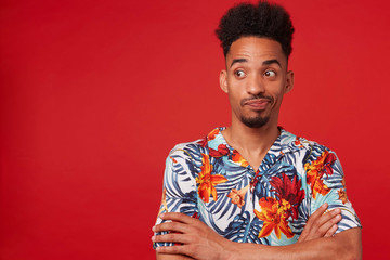 Wall Mural - Young unhappy African American guy wears in Hawaiian shirt, looking left at the copy space, stands over red background with crossed arms.