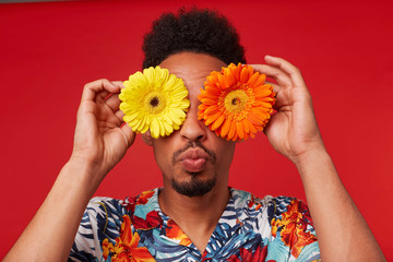 Wall Mural - Close up positive young African American guy, wears in Hawaiian shirt, looks at the camera trough flowers with happy expression, send kiss at the camera, stands over red background.