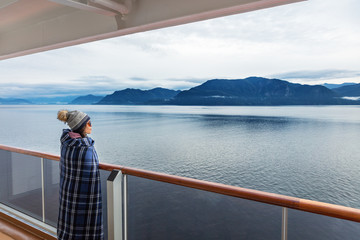 Wall Mural - Alaska cruise travel luxury vacation woman watching inside passage scenic cruising day on balcony deck enjoying view of mountains and nature landscape. Asian girl tourist with wool blanket.