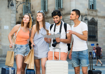 four cheerful travelers holding map in hands and looking for their way in european town